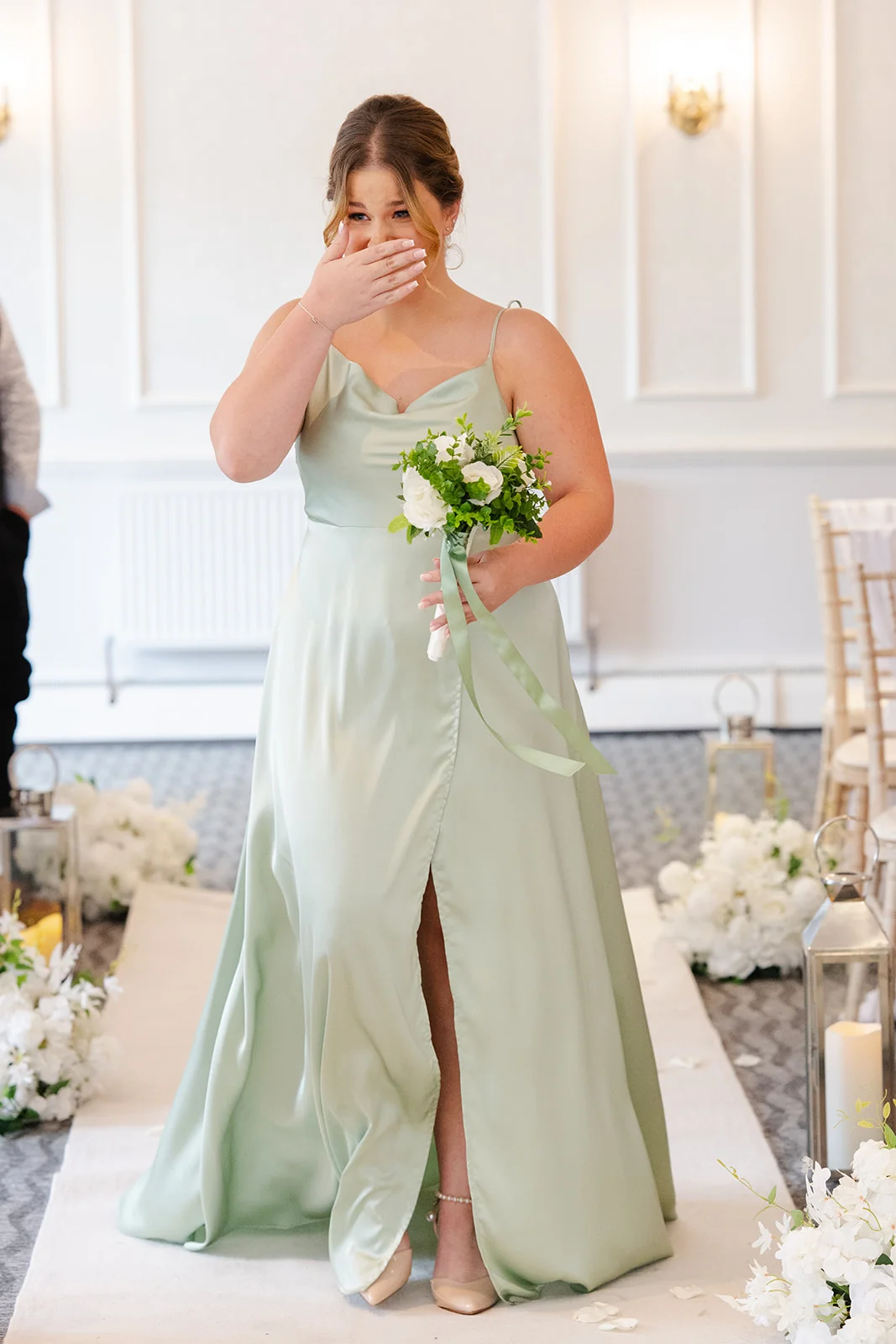An emotional plus size bride walks down the aisle in a sage green satin slip dress with a high split, covering her mouth with her hand as she holds a small bouquet of white roses.