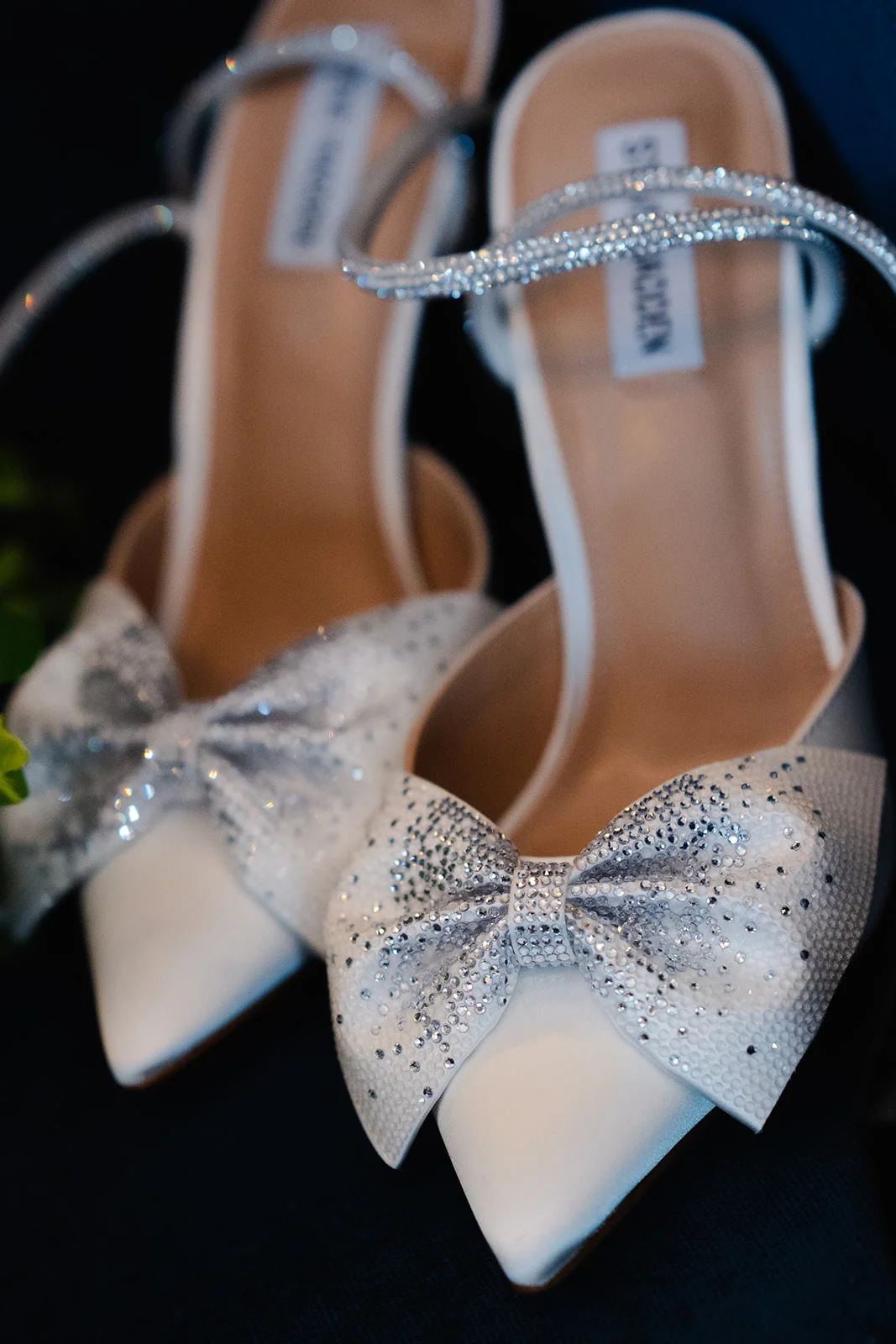 A close-up shot of elegant white satin bridal heels adorned with sparkling crystal bows and embellished ankle straps.
