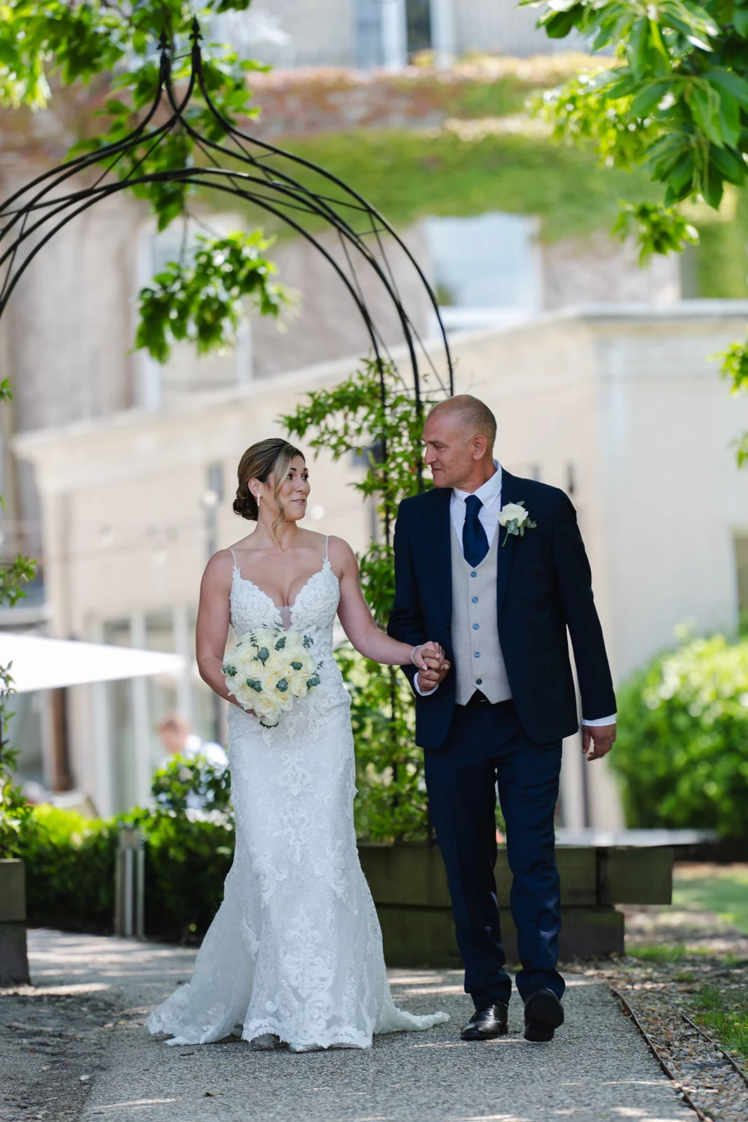 A smiling bride and groom walk hand in hand along a garden path, passing beneath a black metal archway on a bright summer day.