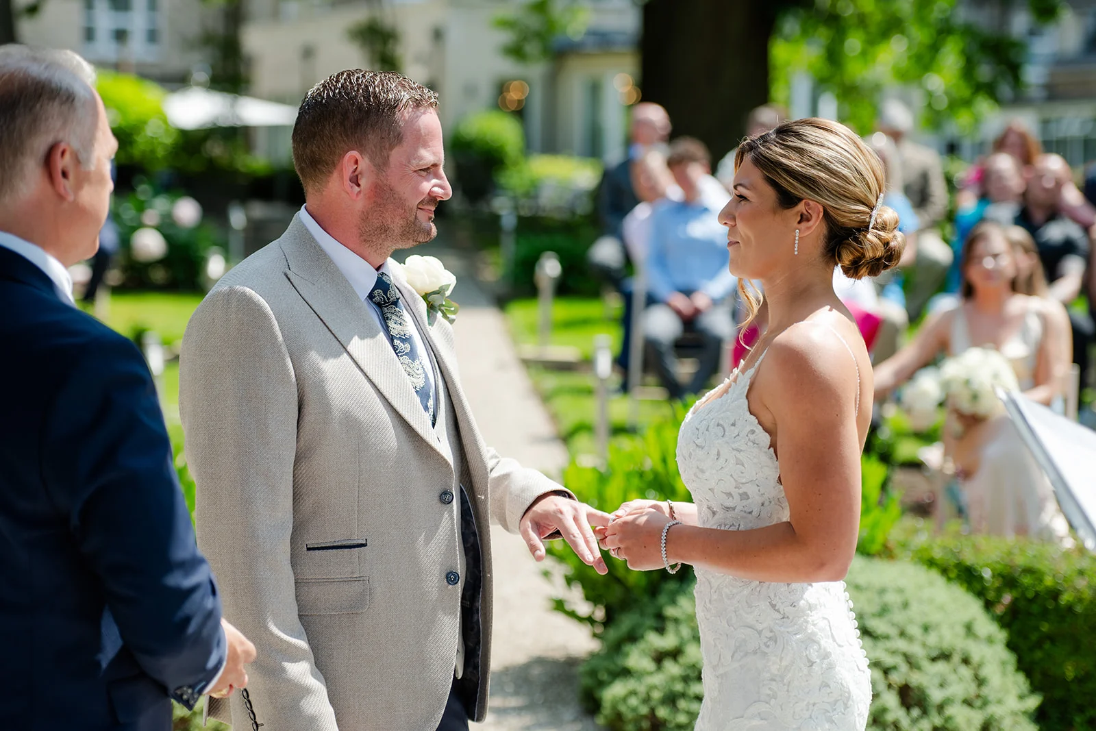 The groom gently slips a wedding ring onto the bride's finger during a sunlit outdoor garden ceremony.