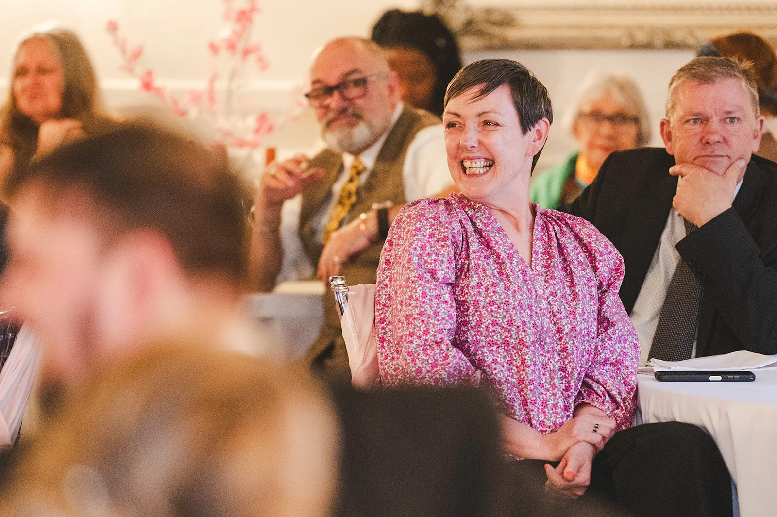 A guest wearing a pink floral blouse laughs joyfully during the wedding speeches, surrounded by other attentive guests.