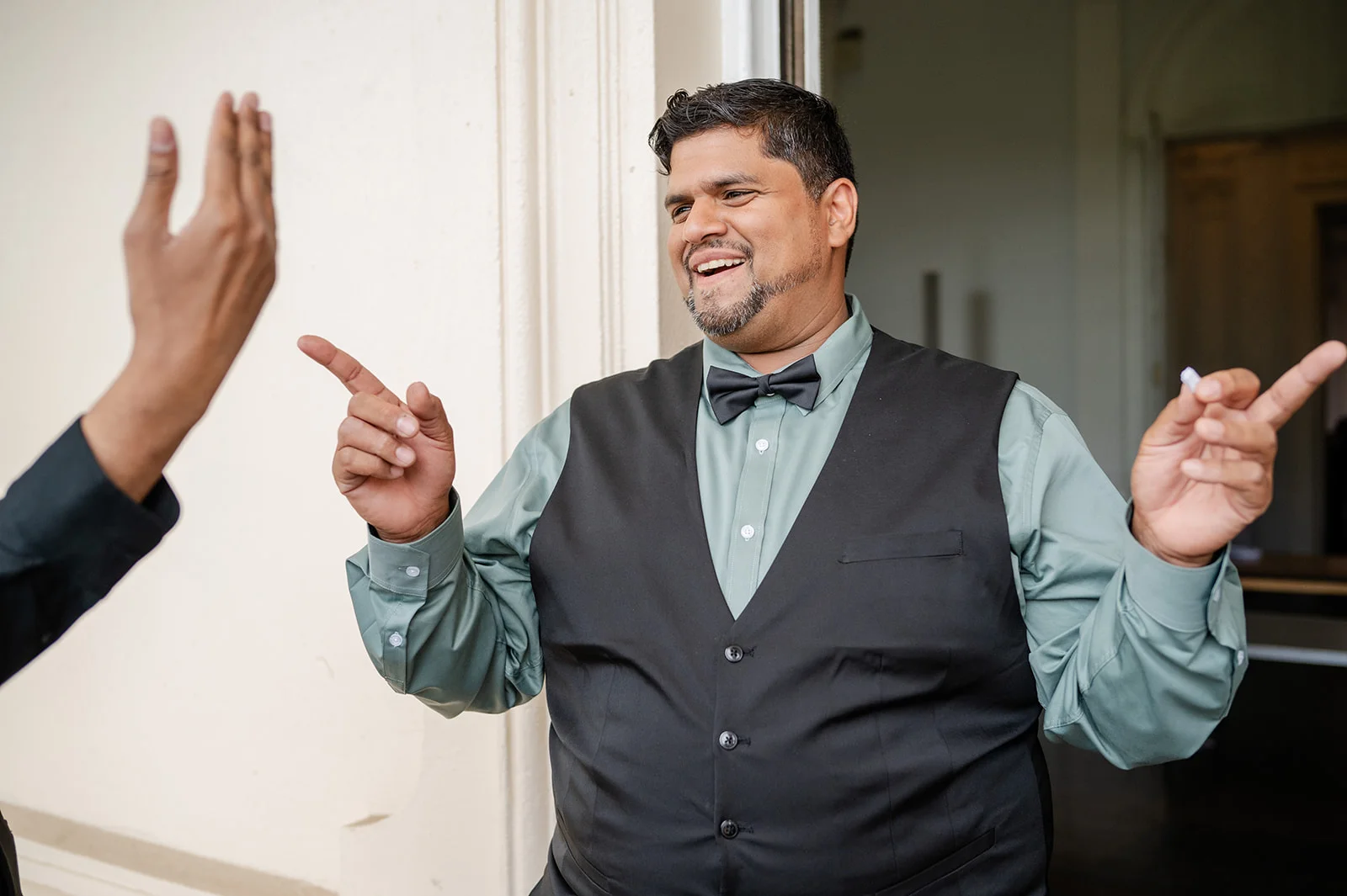 A joyful groom wearing a sage shirt, black waistcoat and bow tie laughs playfully while gesturing with his hands during a candid wedding moment.