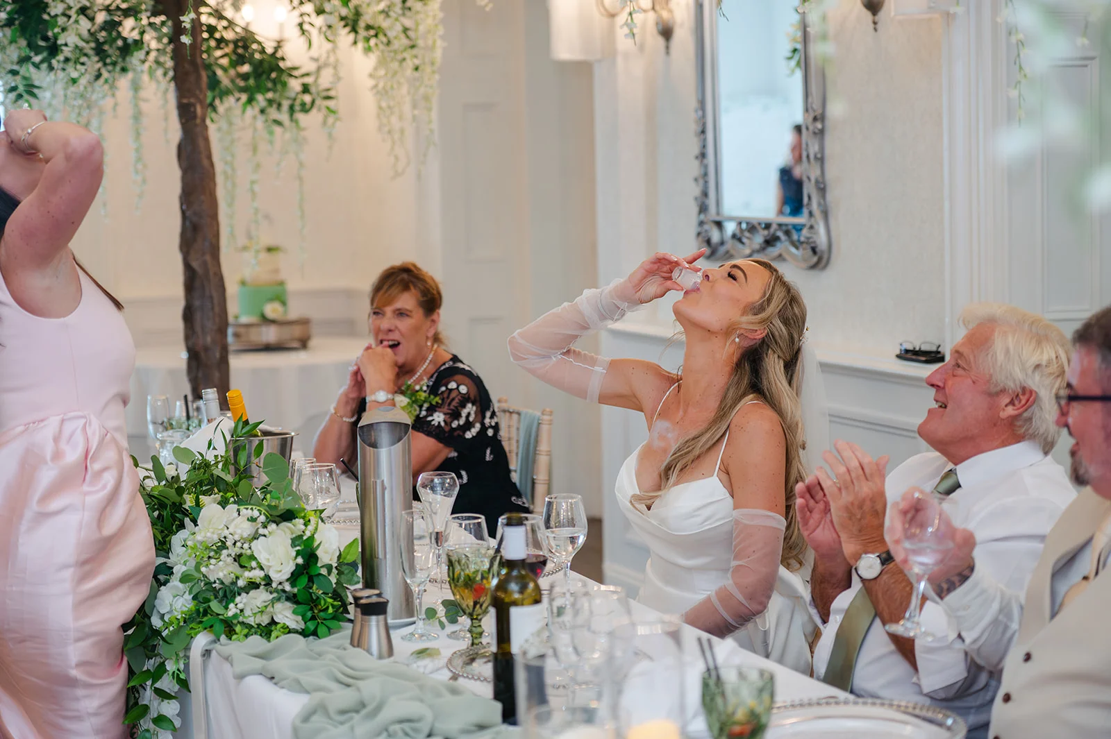 A joyful bride in a white gown and sheer sleeves enthusiastically takes a shot at the top table during wedding speeches, surrounded by laughing guests.