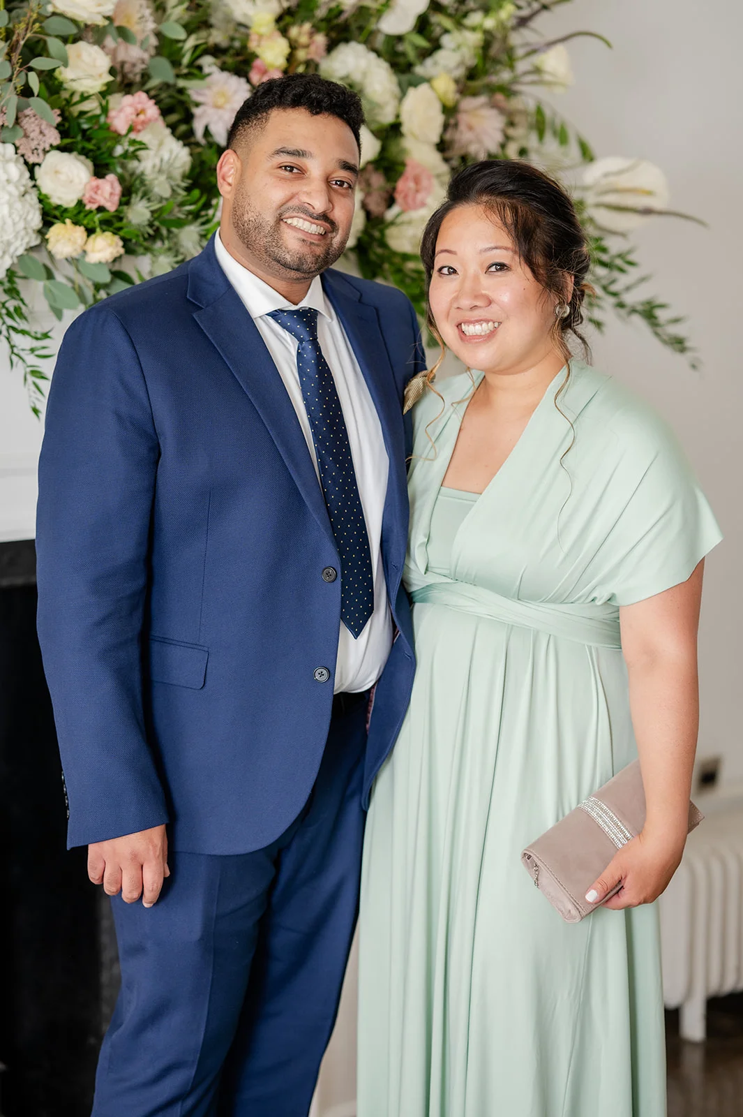 An interracial wedding guest couple smiling together in front of a lush floral archway, with the man in a navy suit and patterned tie and the woman in a sage green dress holding a clutch.