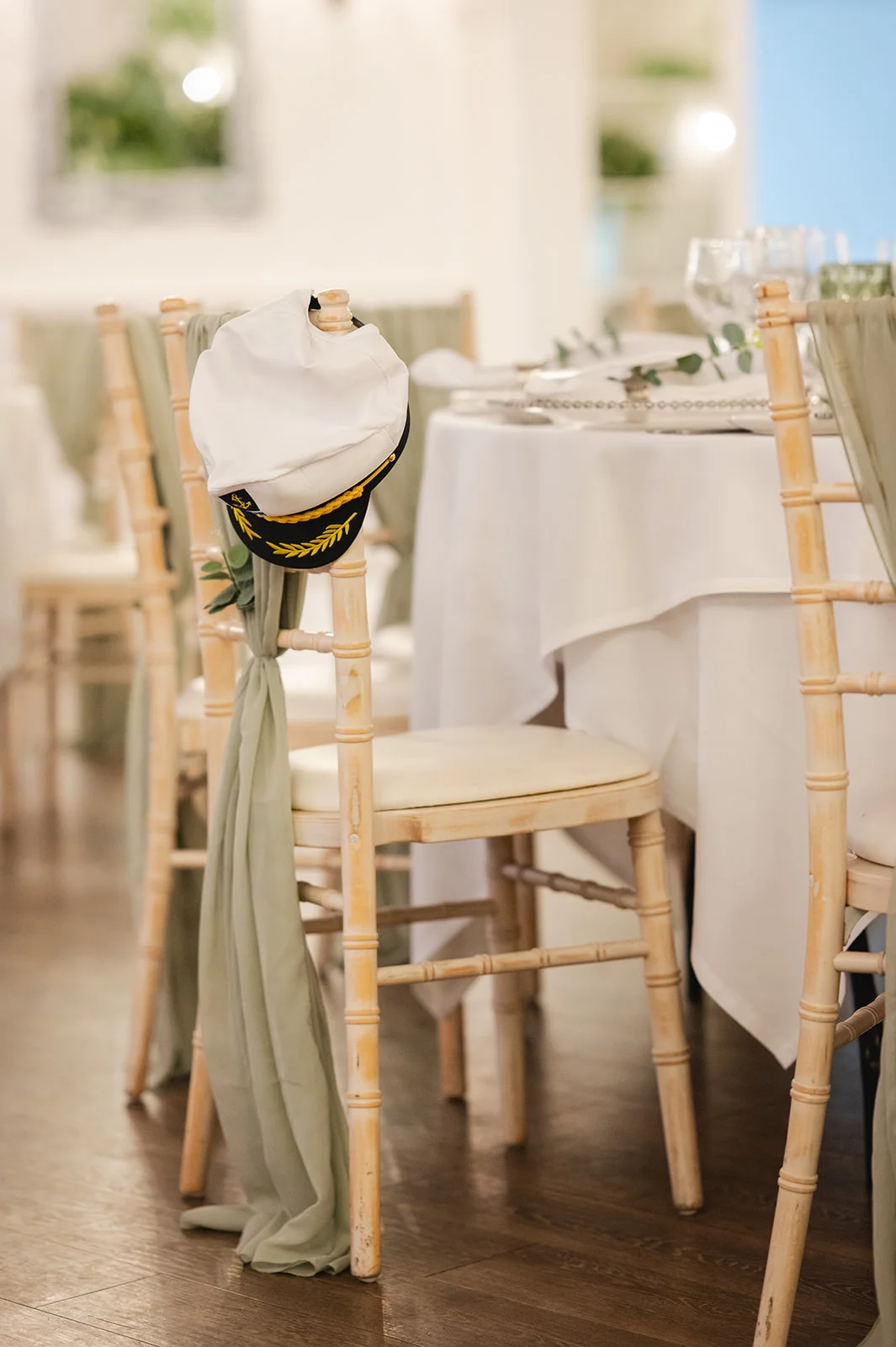 A white captain hat hangs on the back of a wooden chiavari chair adorned with sage green fabric at a wedding reception.