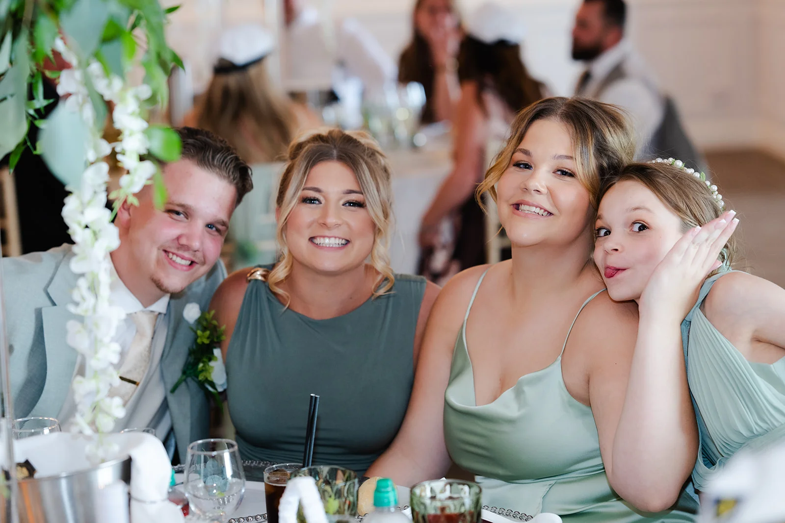 A cheerful group of wedding guests, including bridesmaids in sage green dresses, smiling and posing together at the reception table.