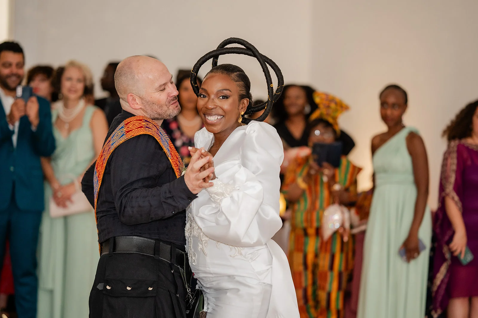 An interracial newlywed couple smiling joyfully while sharing their first dance surrounded by watching wedding guests.