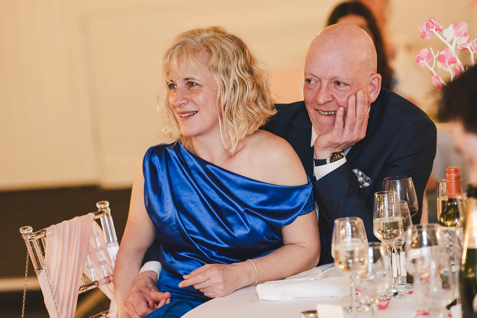 An older couple smiling happily together at a wedding reception table, with the woman wearing a vibrant blue one-shoulder satin gown and the man in a dark suit with his hand on his chin.