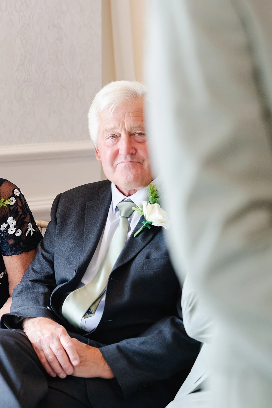 An elderly gentleman with white hair smiles warmly while wearing a dark suit, light tie, and a white floral boutonniere during the wedding celebration.