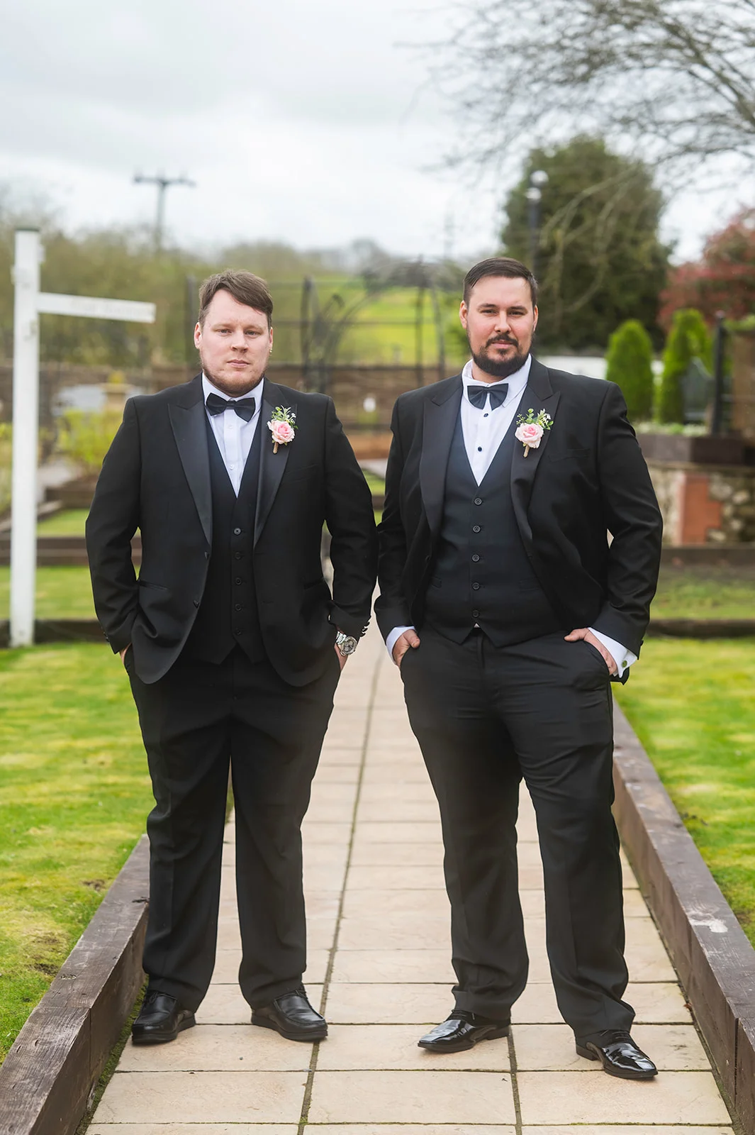 Two grooms in classic black three-piece tuxedos and bow ties pose together on an outdoor stone path framed by green lawns.