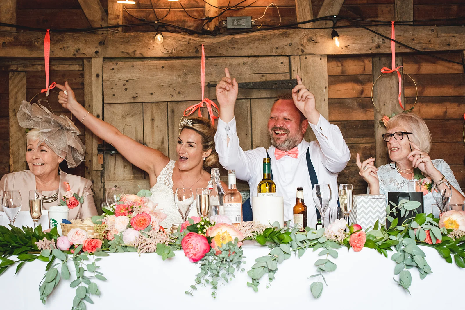 A joyful bride and groom raise their hands in celebration at the top table during wedding speeches inside a rustic wooden barn.