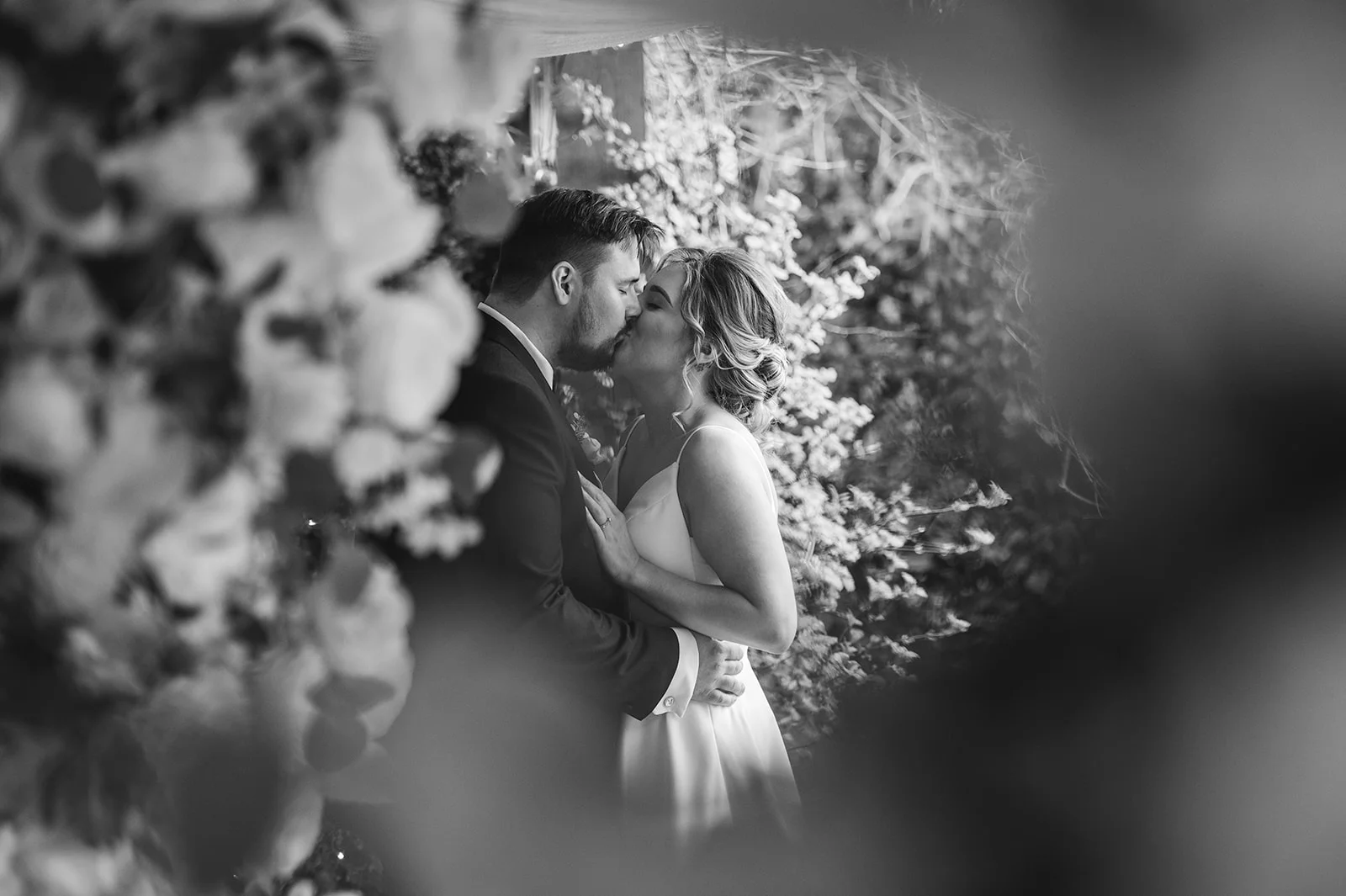 A newlywed couple shares a tender kiss, beautifully framed by lush floral installations and foliage in a romantic black and white photograph.