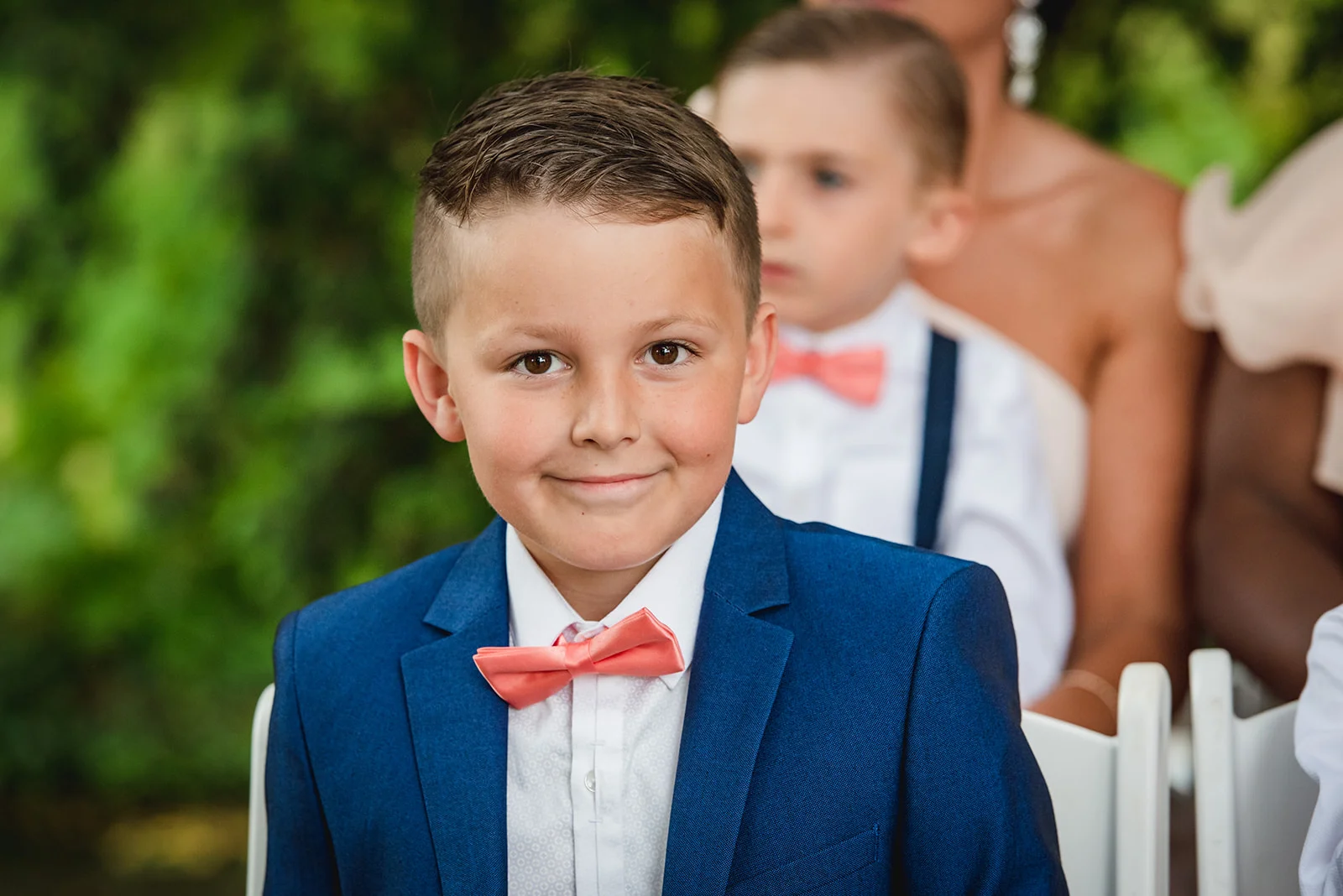 A young page boy wearing a smart navy blue suit and a peach-coloured bow tie smiles gently during a wedding ceremony.