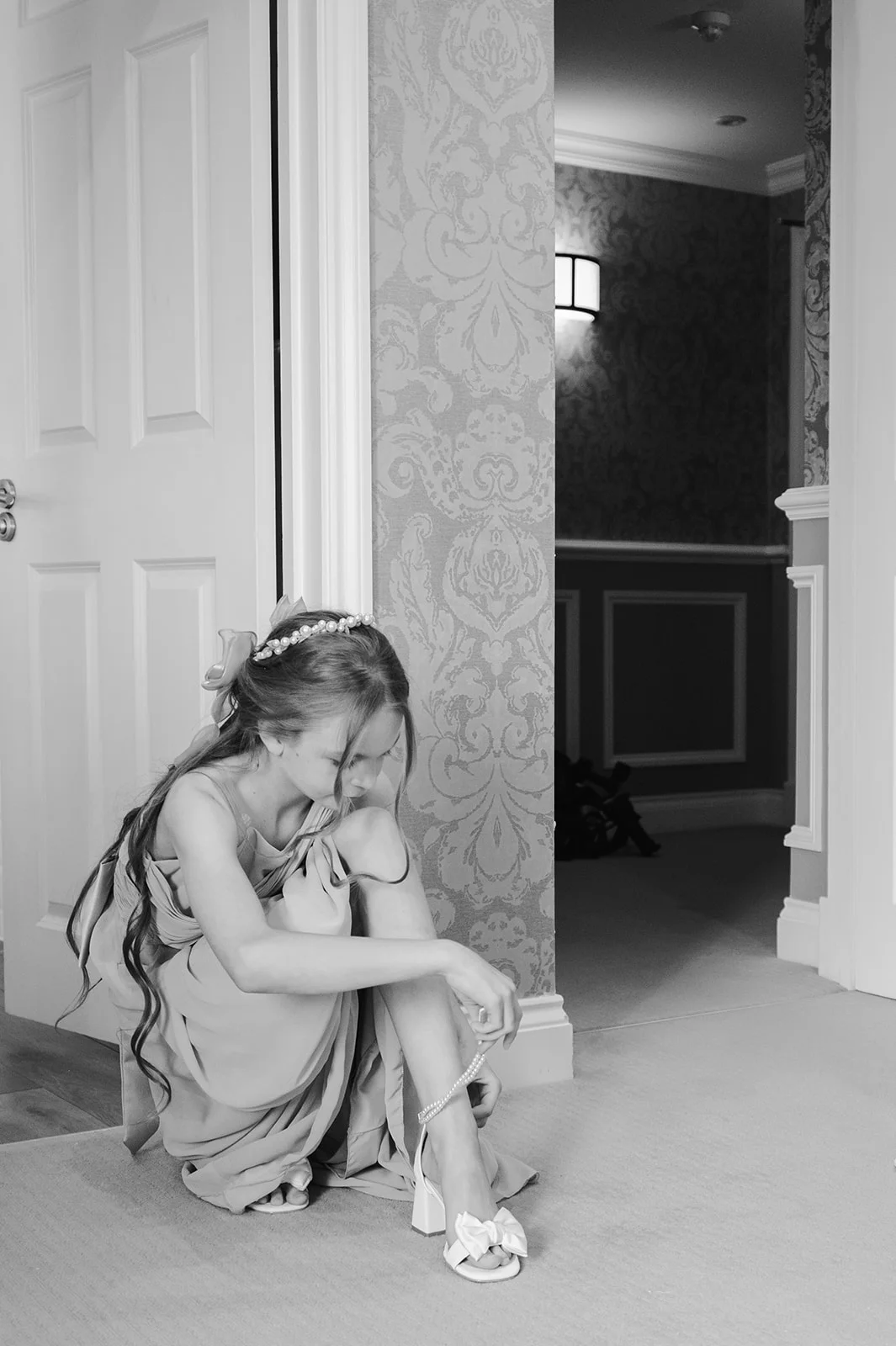 A young flower girl in a formal dress sits on the floor near an open doorway, carefully fastening her block-heeled bow shoe.