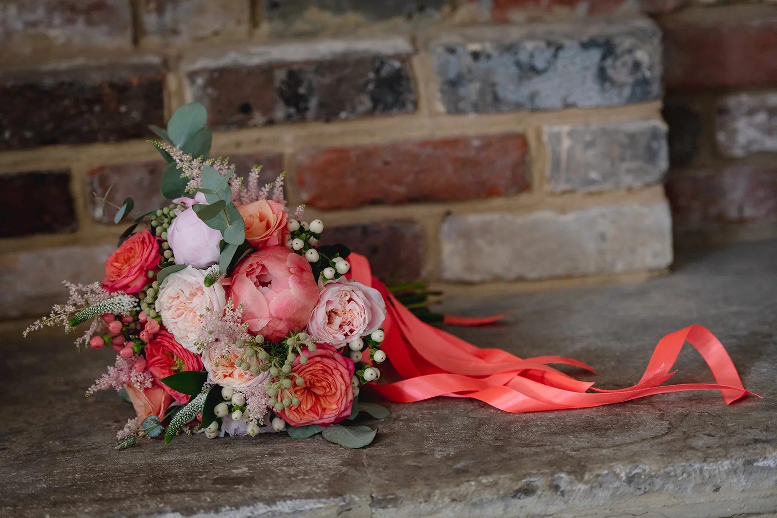 A lush coral and pink bridal bouquet with trailing ribbon rests on a stone surface against an exposed brick wall.