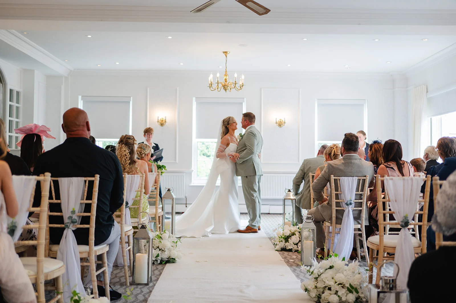 A bride in a white gown and a groom in a sage suit embrace at the top of the aisle during an indoor civil ceremony.