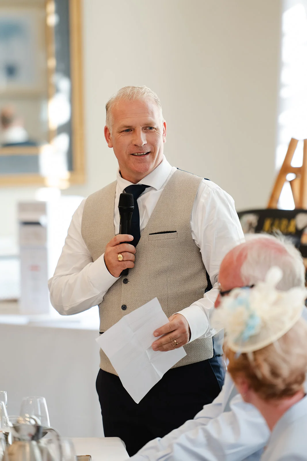 A groom delivers a speech holding a microphone and notes during the wedding reception while guests listen.
