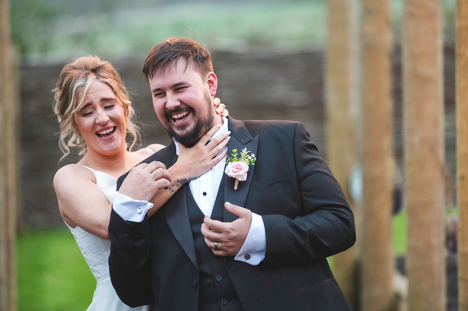 A joyful newlywed bride and groom share a hilarious moment of laughter together outdoors, with the groom wearing a black tuxedo and the bride in a white gown.