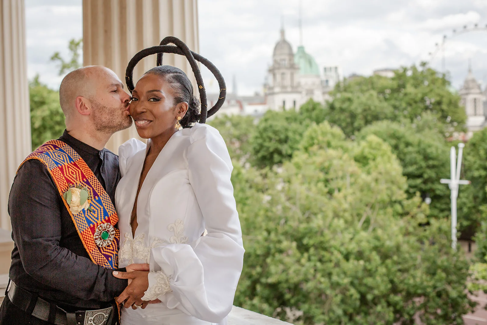 A groom kisses a smiling bride on the cheek against a scenic London city skyline backdrop during an outdoor wedding celebration.