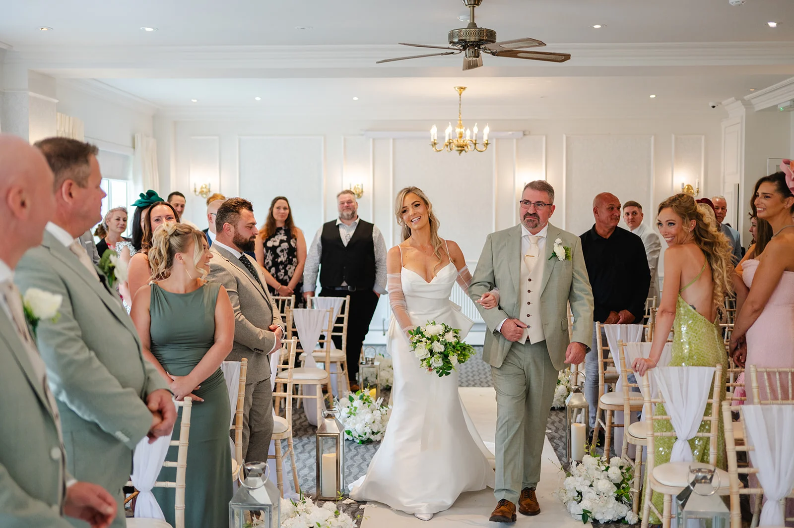 A smiling bride walks down the aisle arm in arm with her father during an indoor wedding ceremony, surrounded by seated guests and elegant floral arrangements.