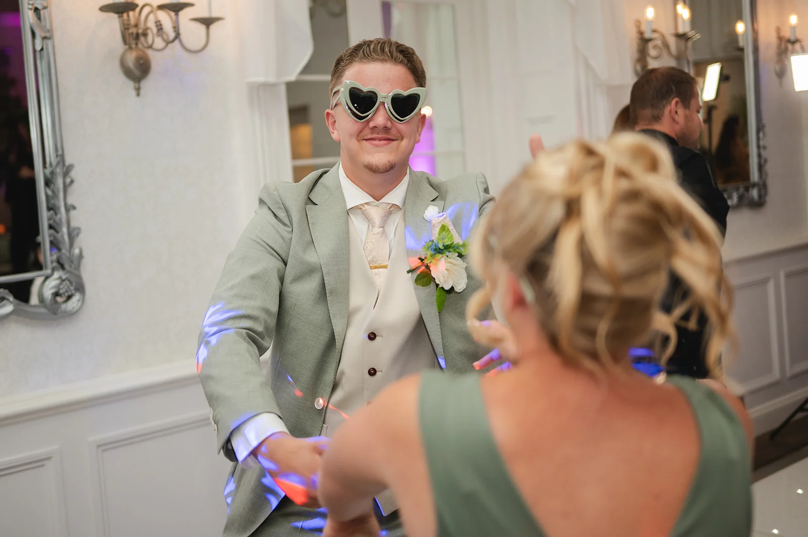 A groom wearing oversized heart-shaped sunglasses and a sage green suit enjoys dancing with a guest on the illuminated dance floor.