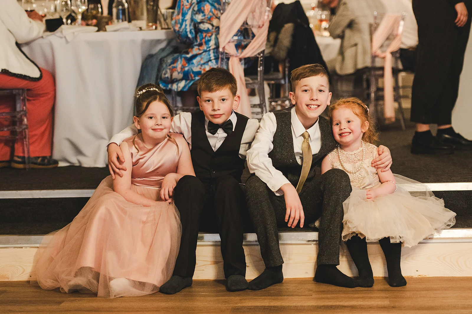 Four children in wedding attire including a flower girl in a pink dress and a page boy in a tweed waistcoat sit happily together on steps during the reception.