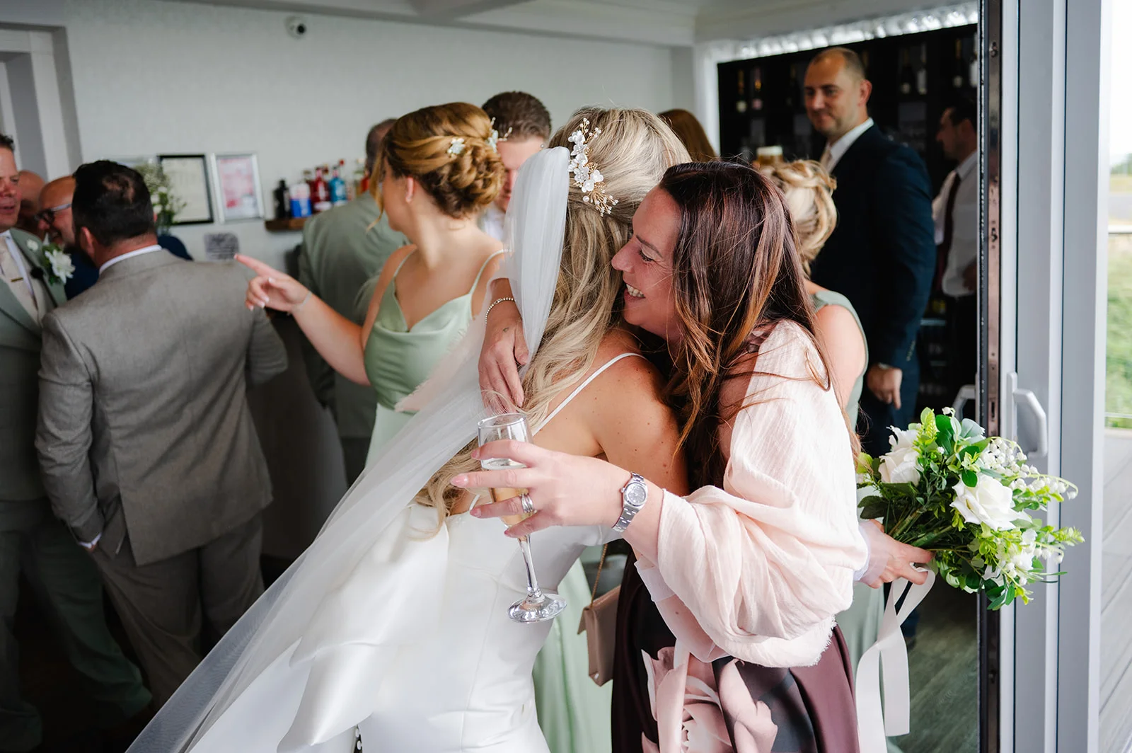 A smiling bride wearing a satin gown and veil embraces a guest warmly while holding a glass of champagne during the wedding reception.