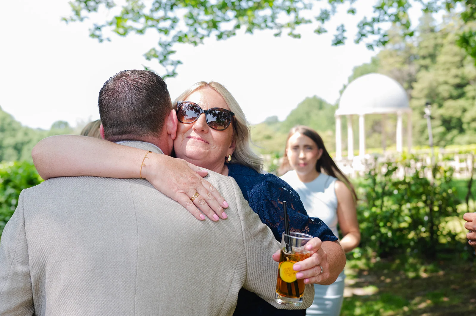 A mother and guest share a warm, emotional embrace outdoors during a sunny wedding reception while holding a refreshing drink.