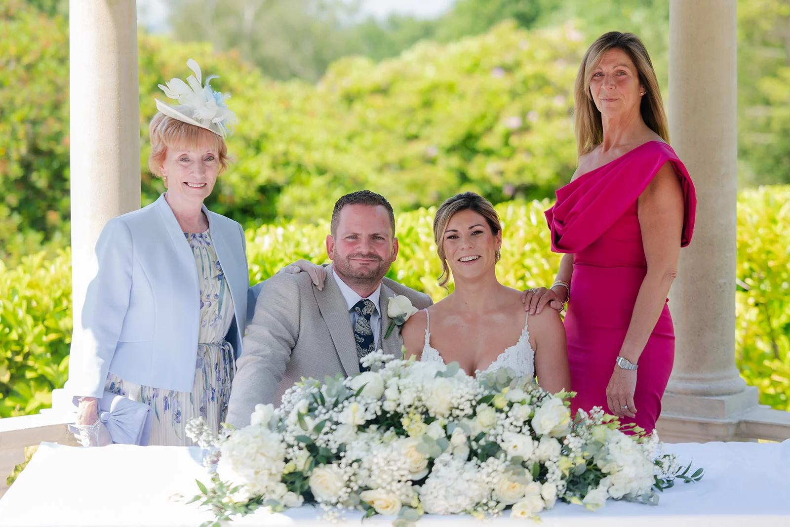 A newlywed couple poses alongside family members beneath a stone gazebo featuring a lush white floral arrangement on the table.