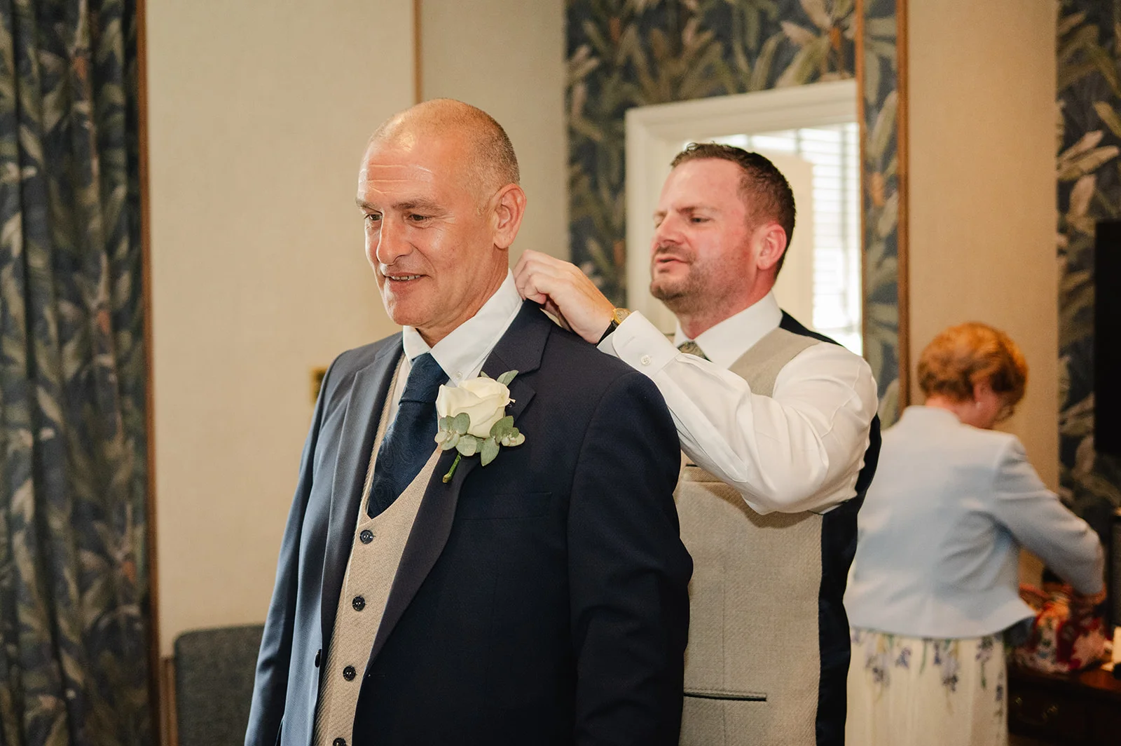 A groom smiling as a groomsman adjusts his tie during morning preparations inside a well-lit room.
