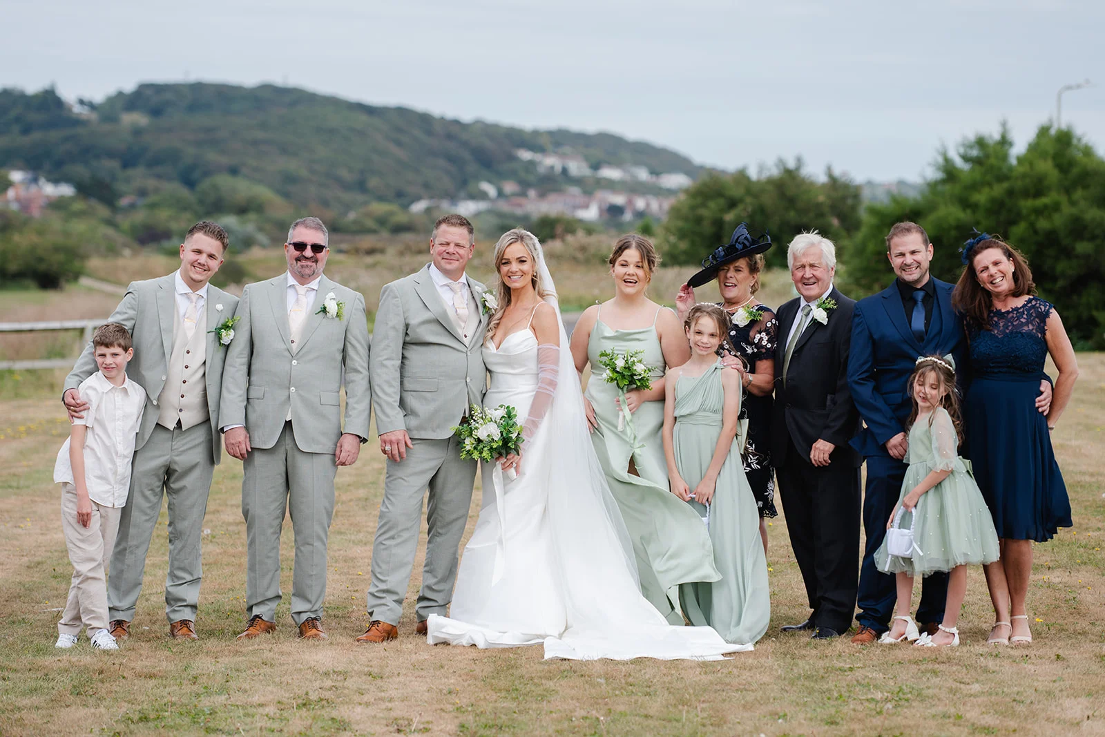 A large wedding party and family group pose outdoors on a grassy field with rolling hills in the background under a soft overcast sky.