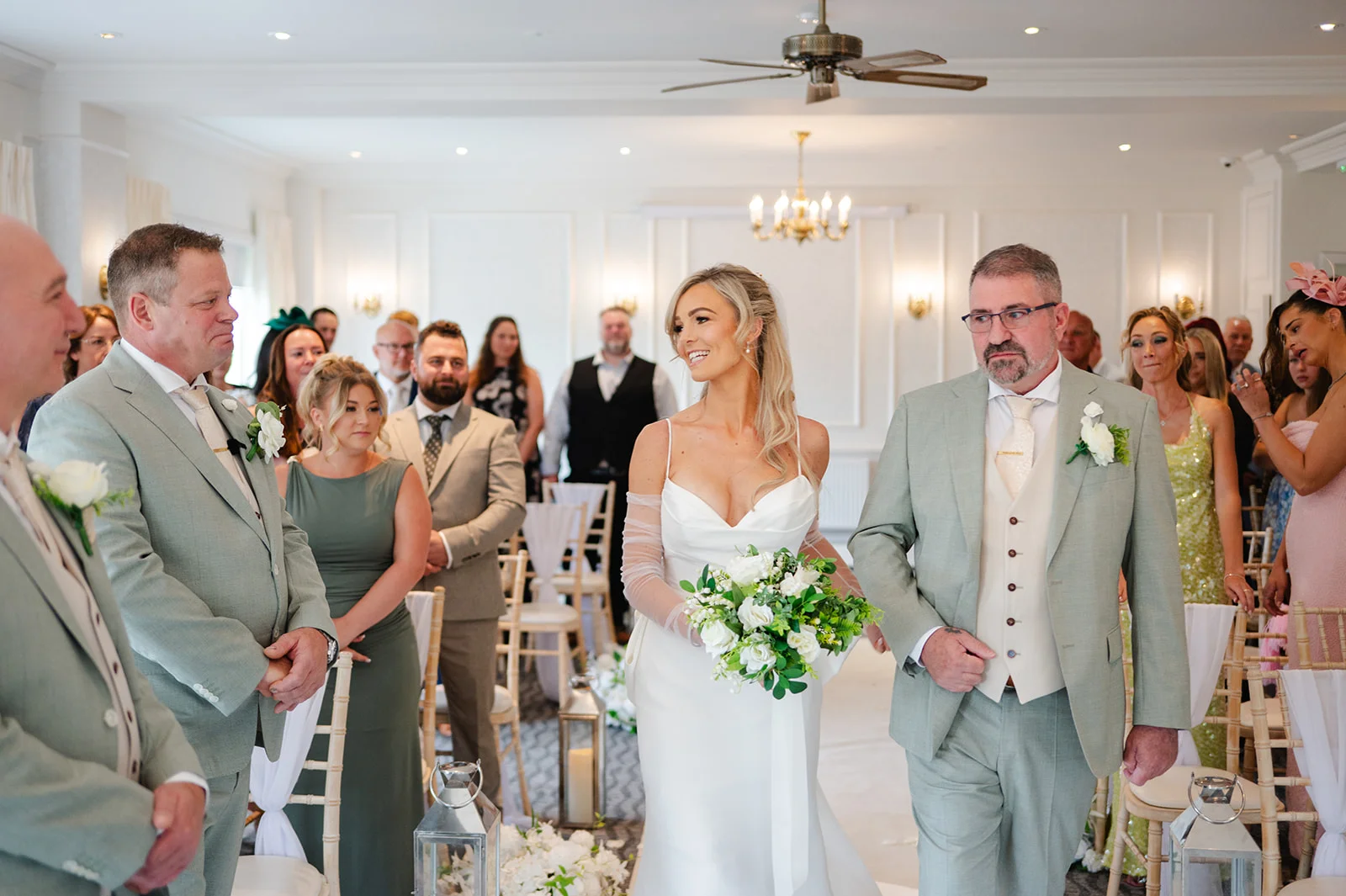 A smiling bride walks down the aisle accompanied by her father, holding a bouquet of white roses and greenery during a civil ceremony.