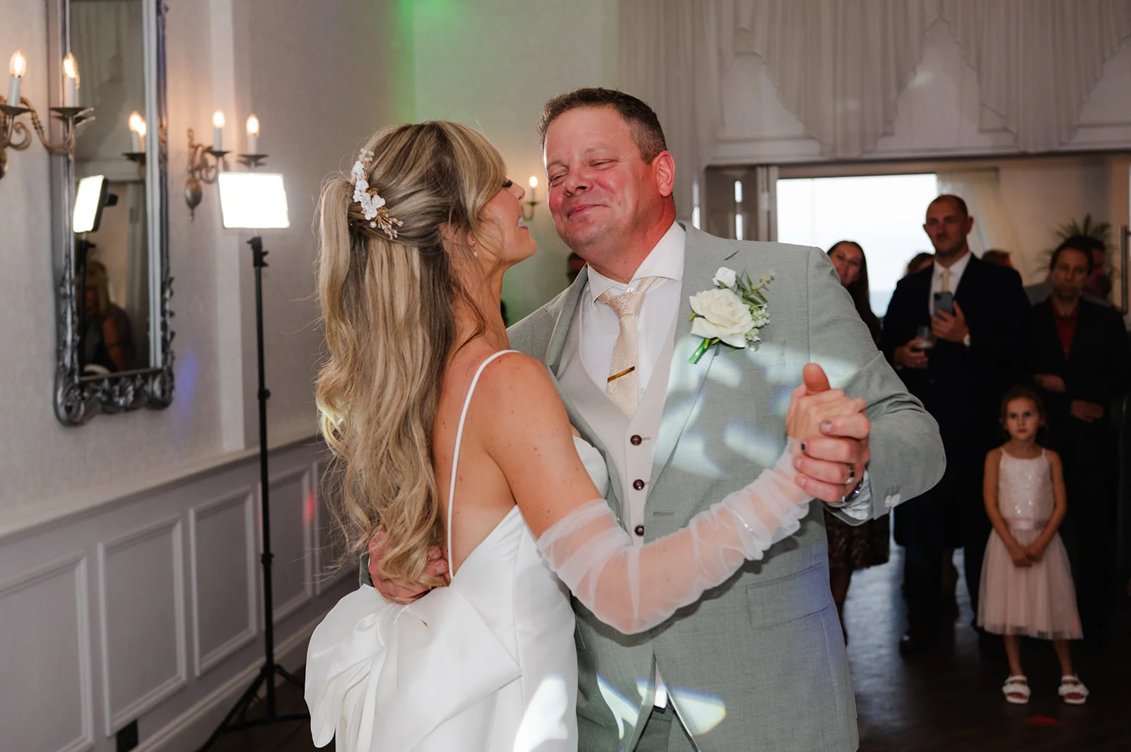The bride and groom share a tender moment during their first dance in a hotel ballroom, surrounded by admiring family and guests.