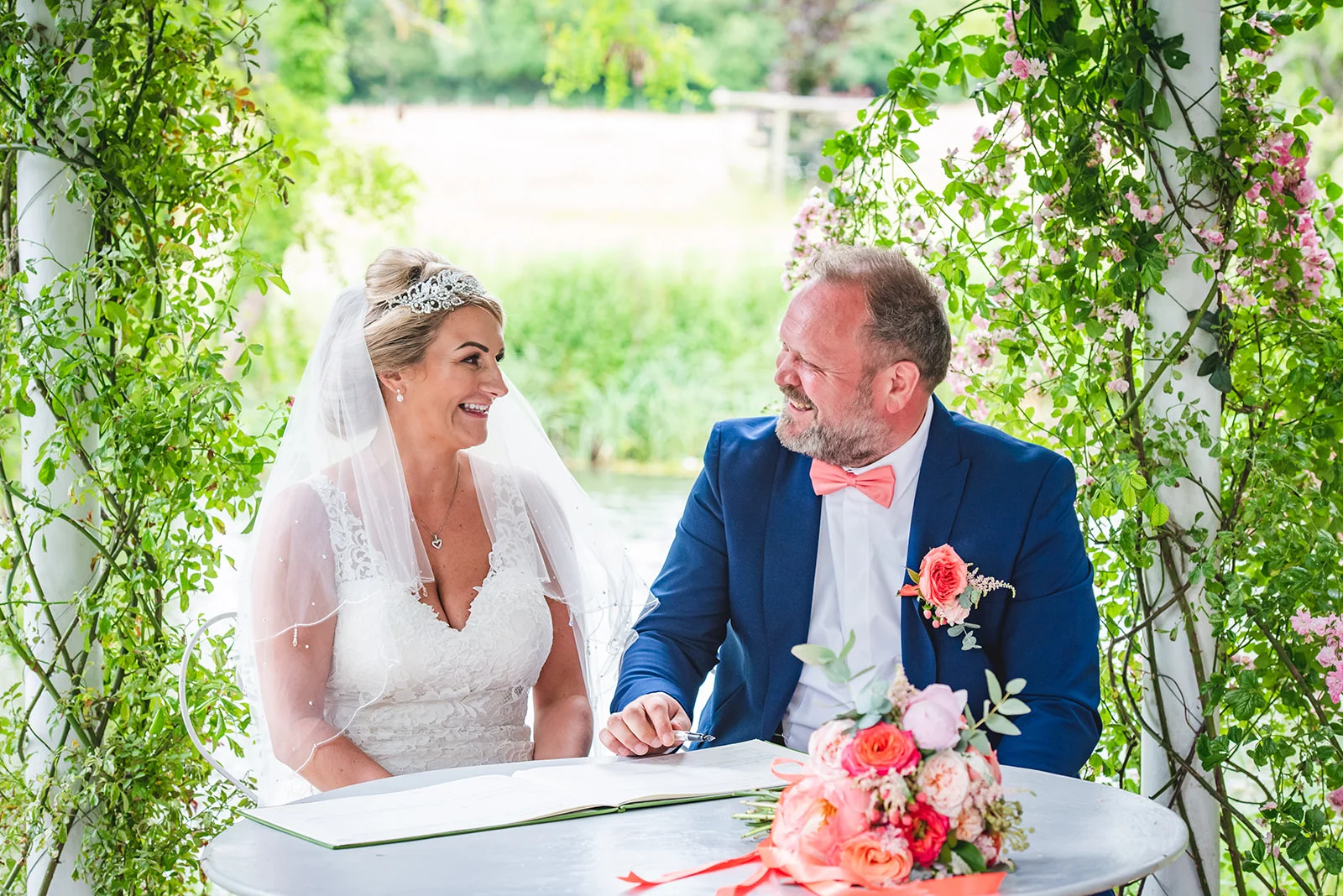 A smiling bride and groom sit together signing the wedding register inside a floral-adorned gazebo during an outdoor summer ceremony.