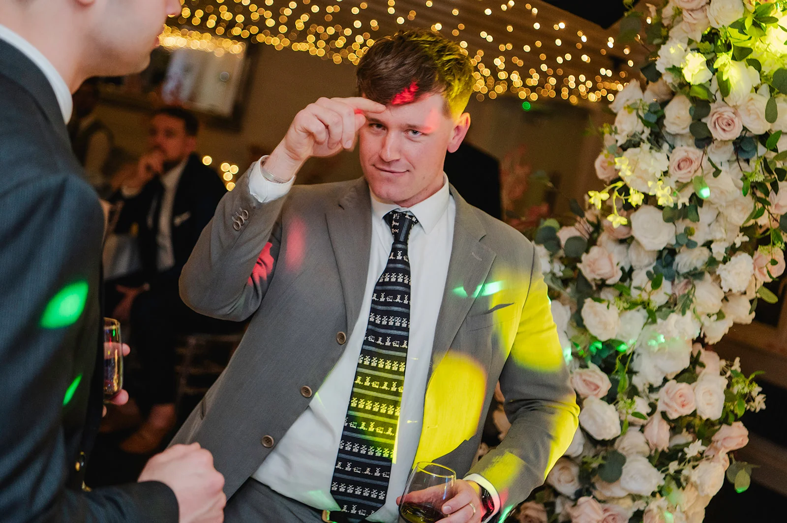 A guest in a grey suit and patterned tie enjoys the evening reception near a large white floral arch while colourful disco lights sweep across the scene.