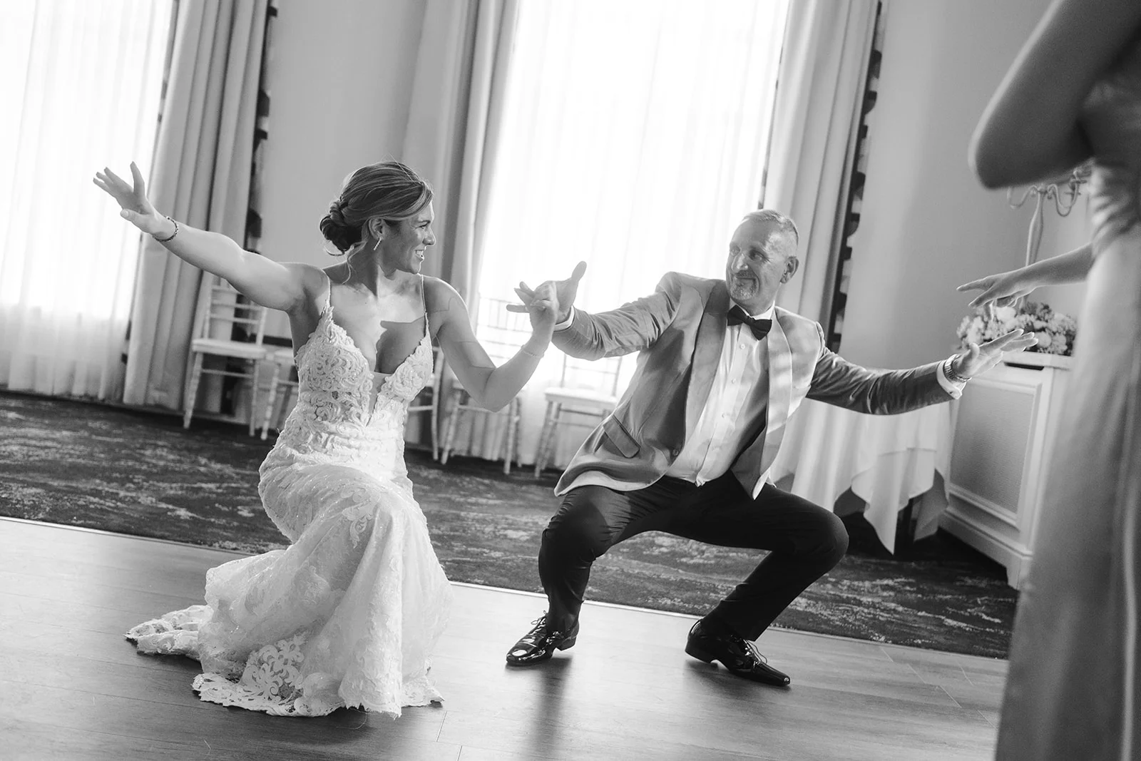 A joyful bride in a lace wedding dress and a groom in a tuxedo laugh while performing a playful dance move on the wooden dance floor during their reception.