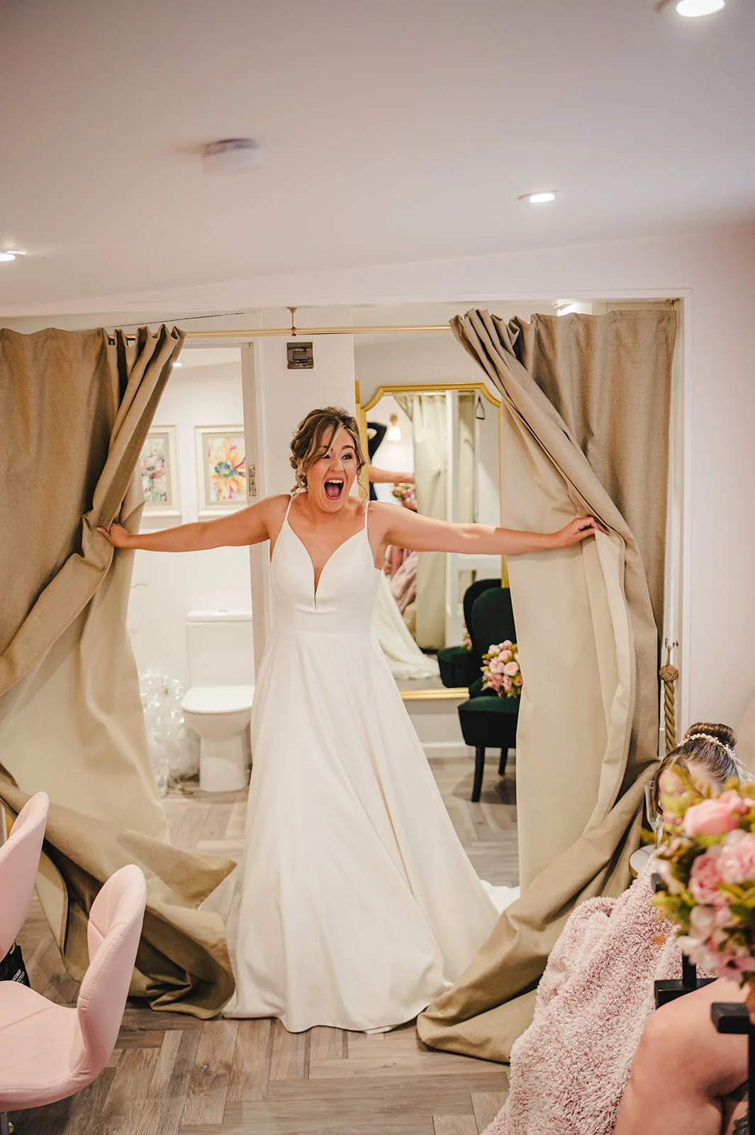 A joyful bride excitedly parts heavy curtains to reveal herself in her white a line wedding dress during morning preparations.