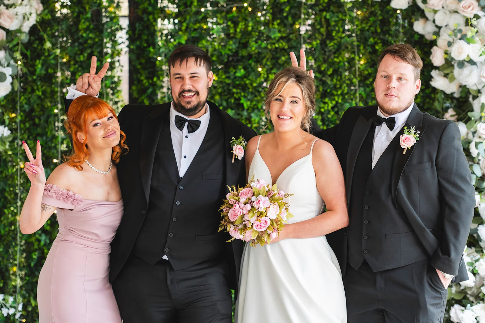 A newly married couple in formal attire pose playfully with their wedding party in front of a lush greenery and floral wall.