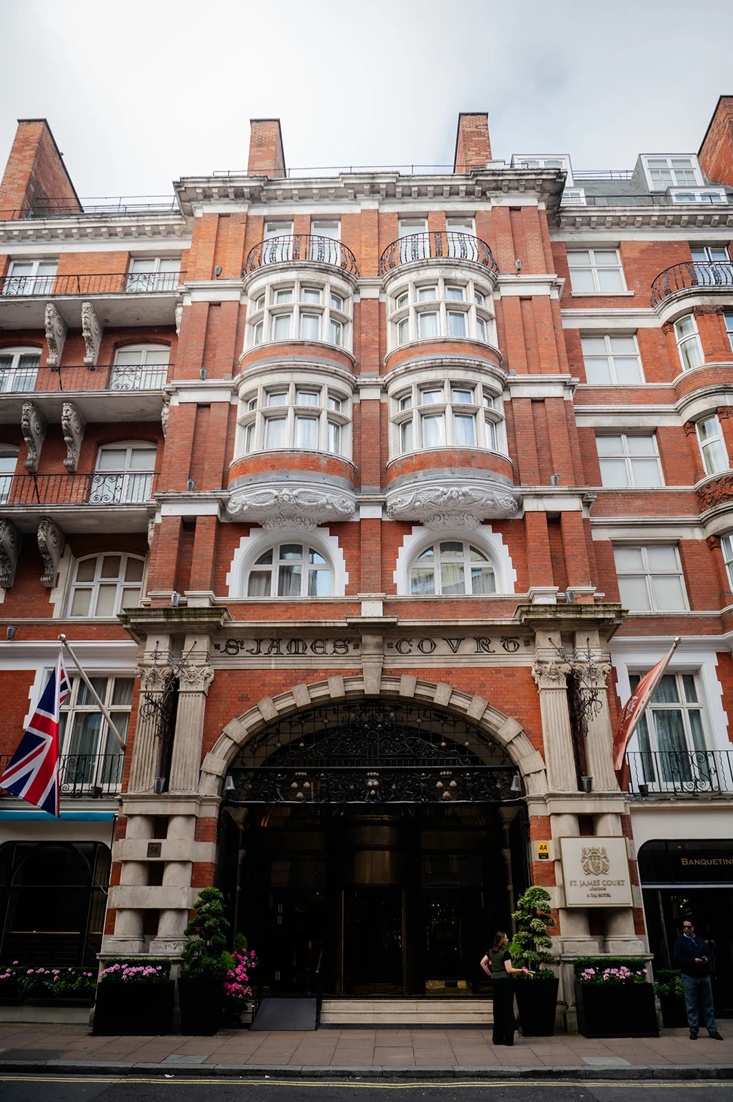 An impressive exterior view of the St. James' Court hotel in London, showcasing a grand red brick facade with arched detailing and a flying Union Jack flag.