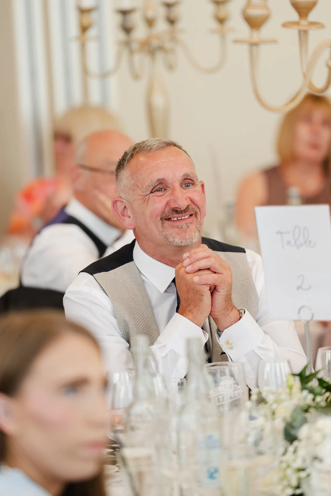 A smiling guest with clasped hands watches wedding speeches at a reception table decorated with floral centrepieces and a table number card.