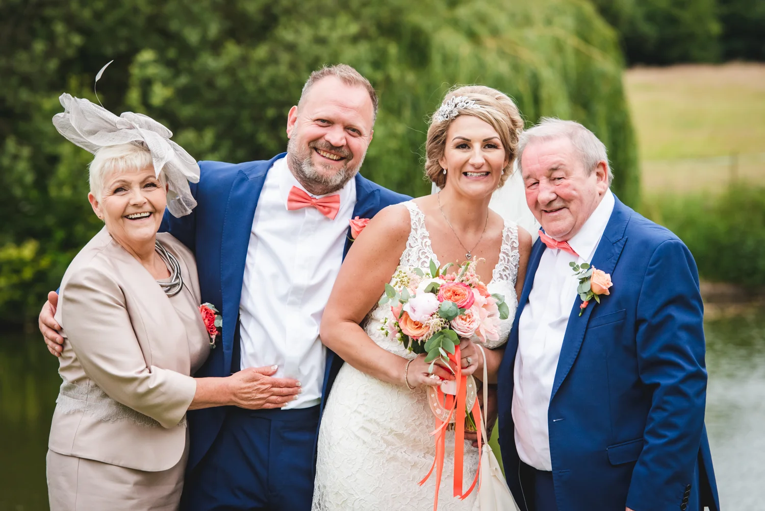 A smiling bride and groom pose happily outdoors alongside the bride's parents near a picturesque lakeside setting during their wedding day.