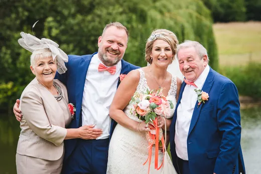 A smiling bride and groom pose happily outdoors alongside the bride's parents near a picturesque lakeside setting during their wedding day.