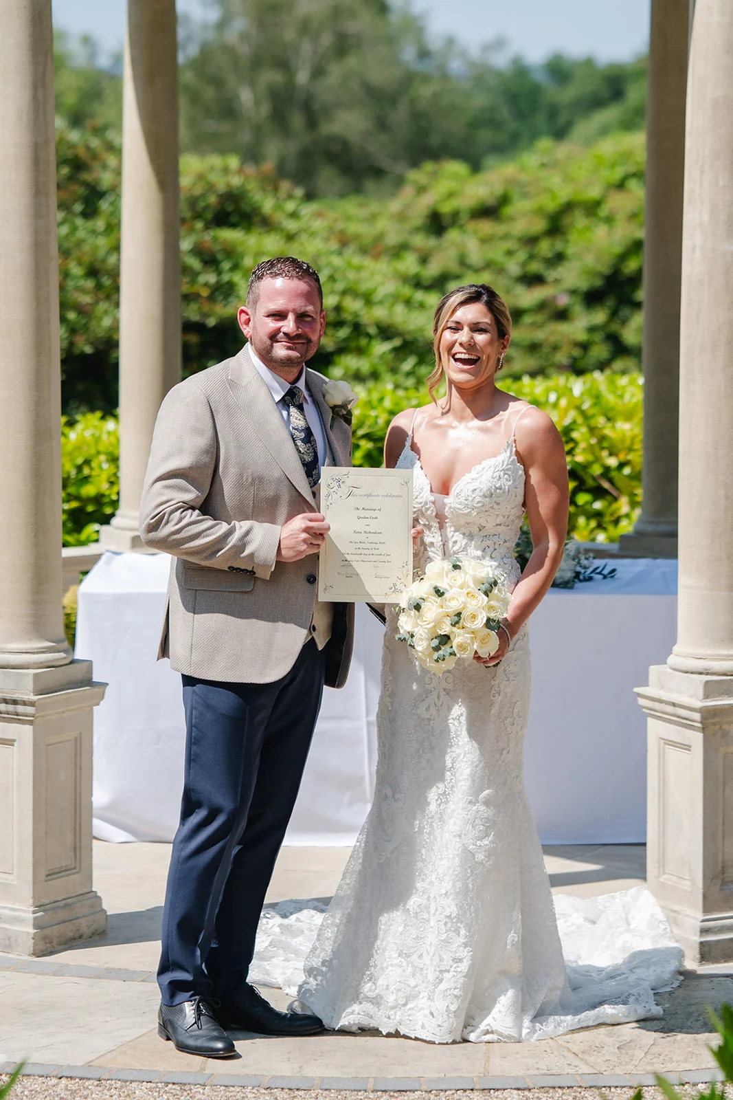 A newlywed couple laugh joyfully while holding their marriage certificate inside an outdoor stone pavilion on a sunny summer wedding day.