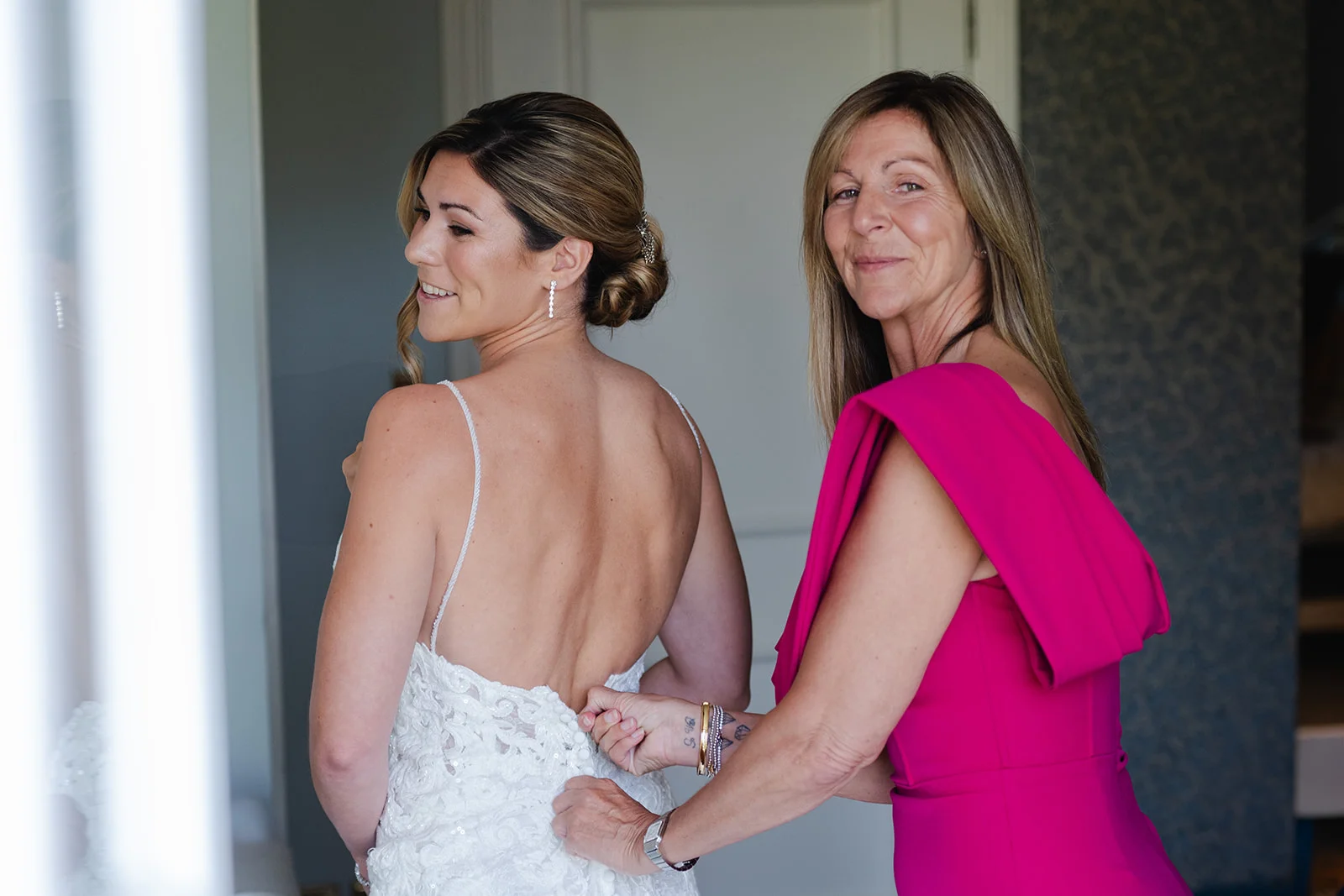 A mother helping her smiling bride daughter zip up her intricate lace wedding dress during bridal preparations.