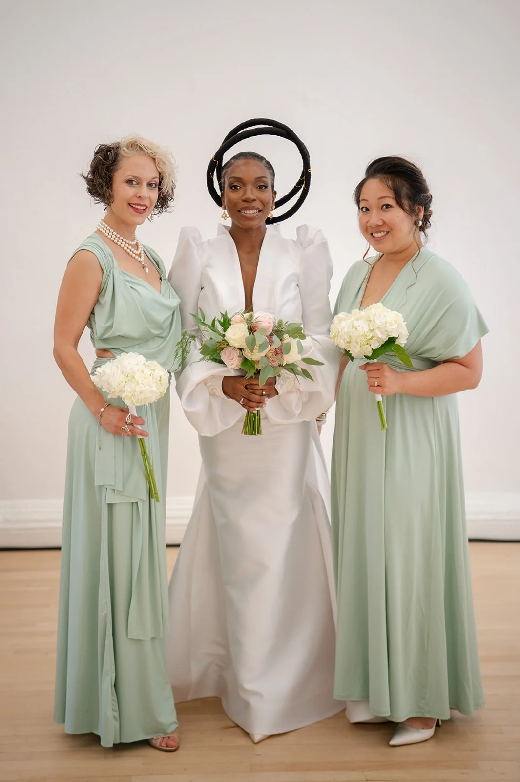 A stylish bride wearing a white structured gown and sculptural halo headpiece poses alongside two bridesmaids in sage green gowns holding bouquets.