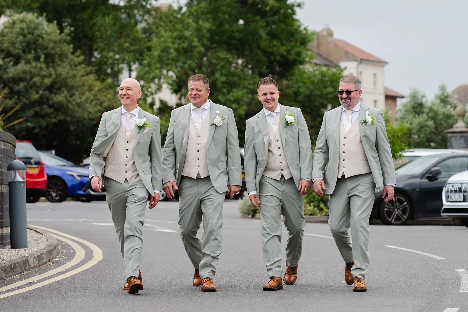 Four smiling groomsmen in matching sage green suits and waistcoats walk together along a paved street on their wedding day.