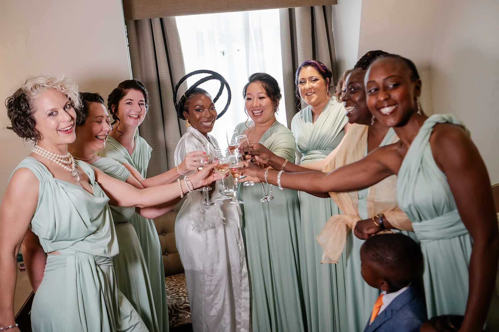 A joyful group of bridesmaids in sage green dresses and a bride wearing a white robe and sculptural headpiece share a celebratory champagne toast while getting ready.