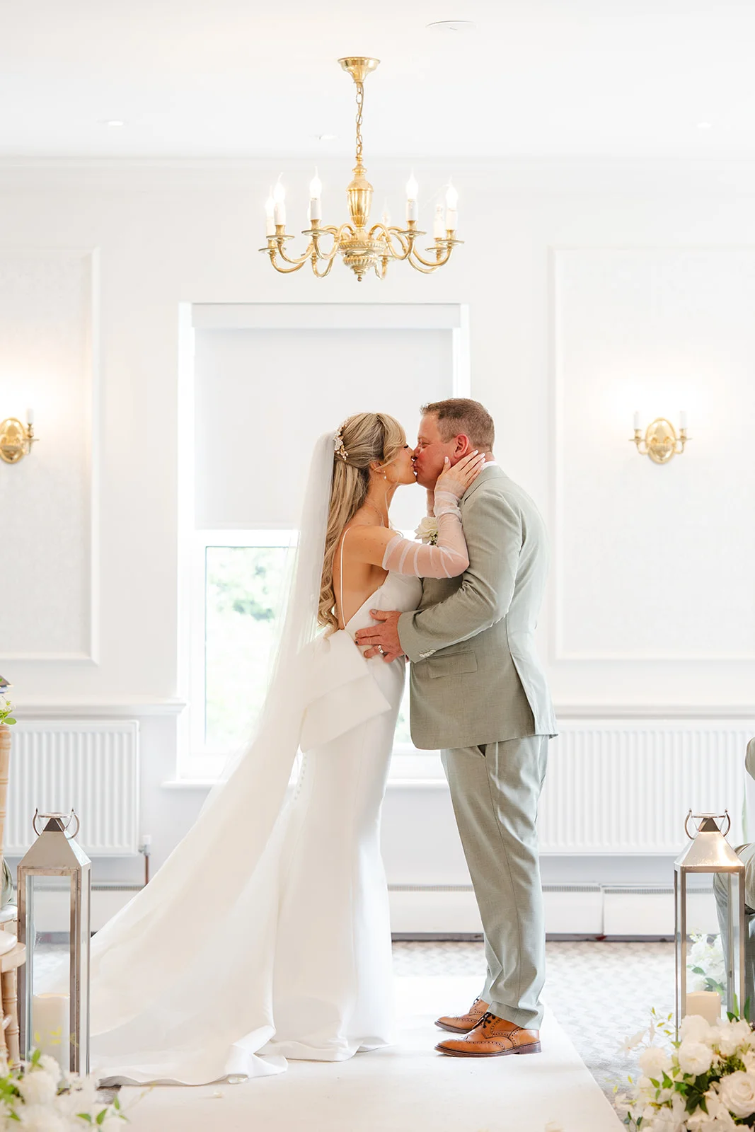 Newlyweds share their first kiss during a bright indoor ceremony inside a light-filled venue featuring a gold chandelier and elegant white decor.