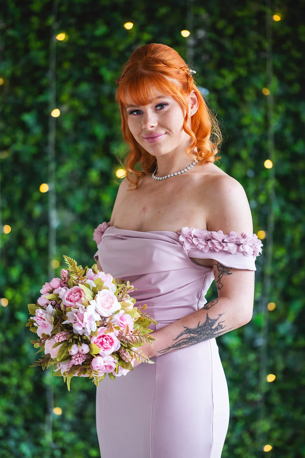 A smiling bridesmaid with vibrant red hair wearing an off-the-shoulder pink dress holds a pink and white floral bouquet in front of a green foliage wall with fairy lights.