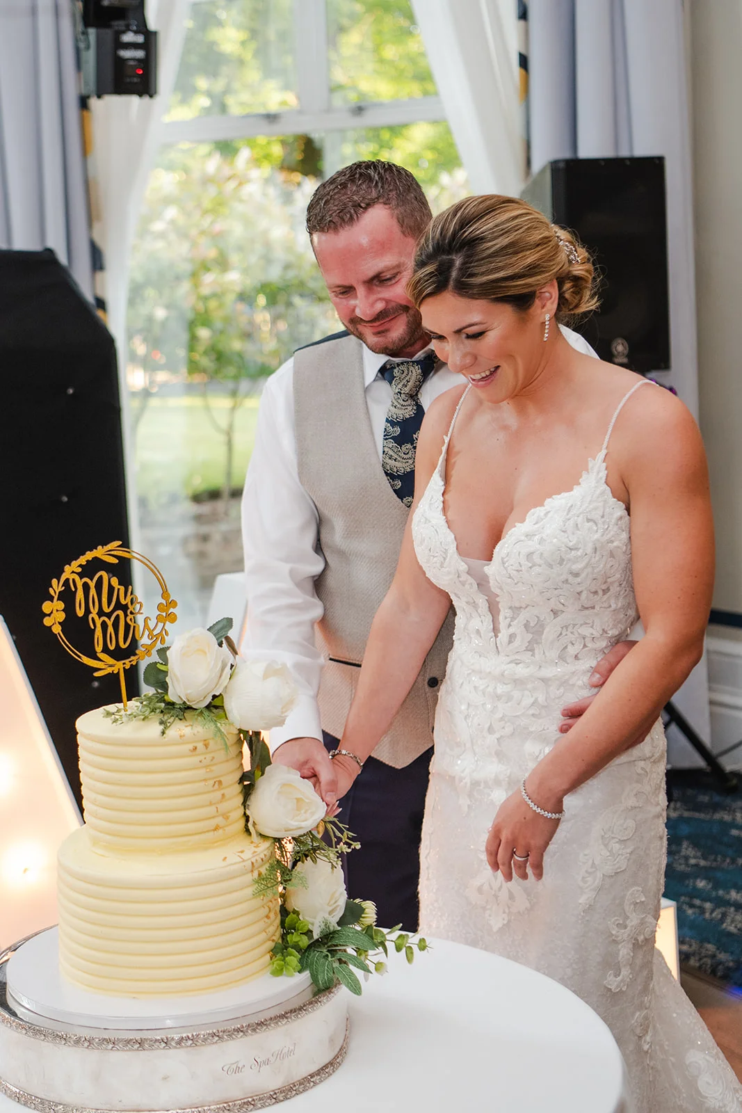 A happy bride and groom laugh together while cutting a two-tiered yellow textured wedding cake adorned with white roses and a gold topper.